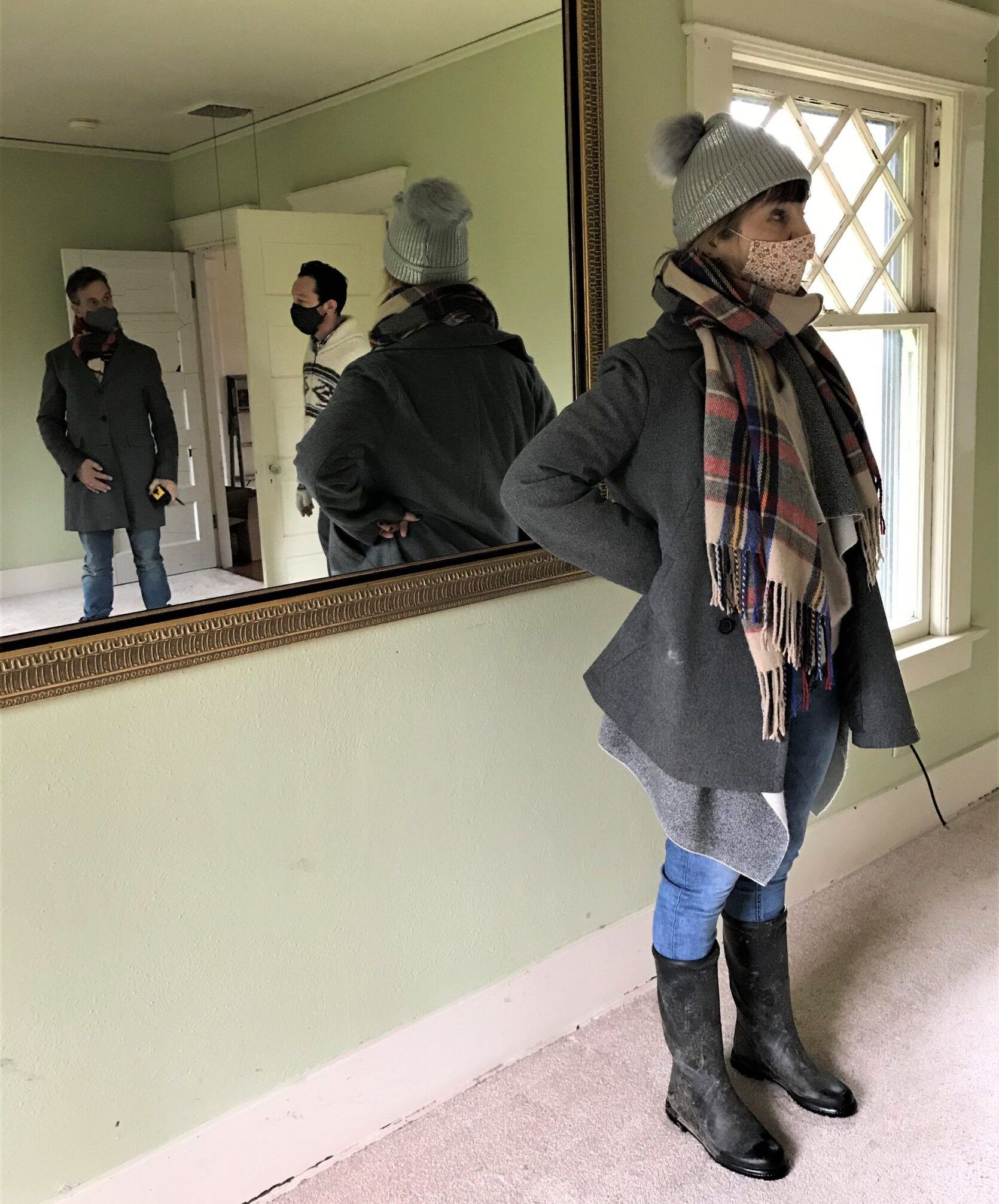 Woman in winter coat stands in room; mirror shows two men in background.