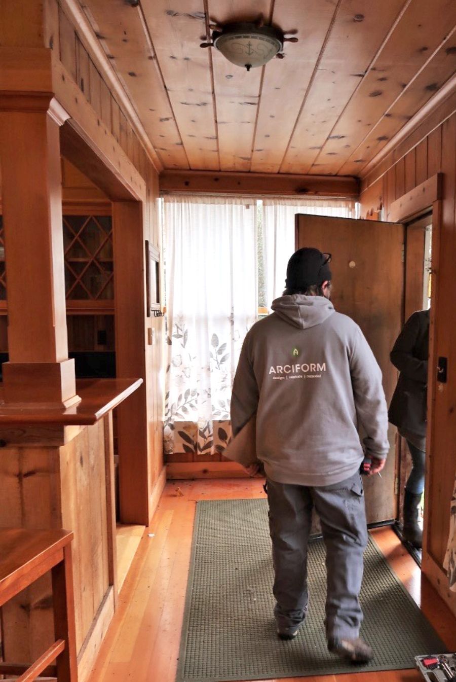 Man in gray hoodie enters a wood-paneled entryway with open door, green rug, and warm lighting.