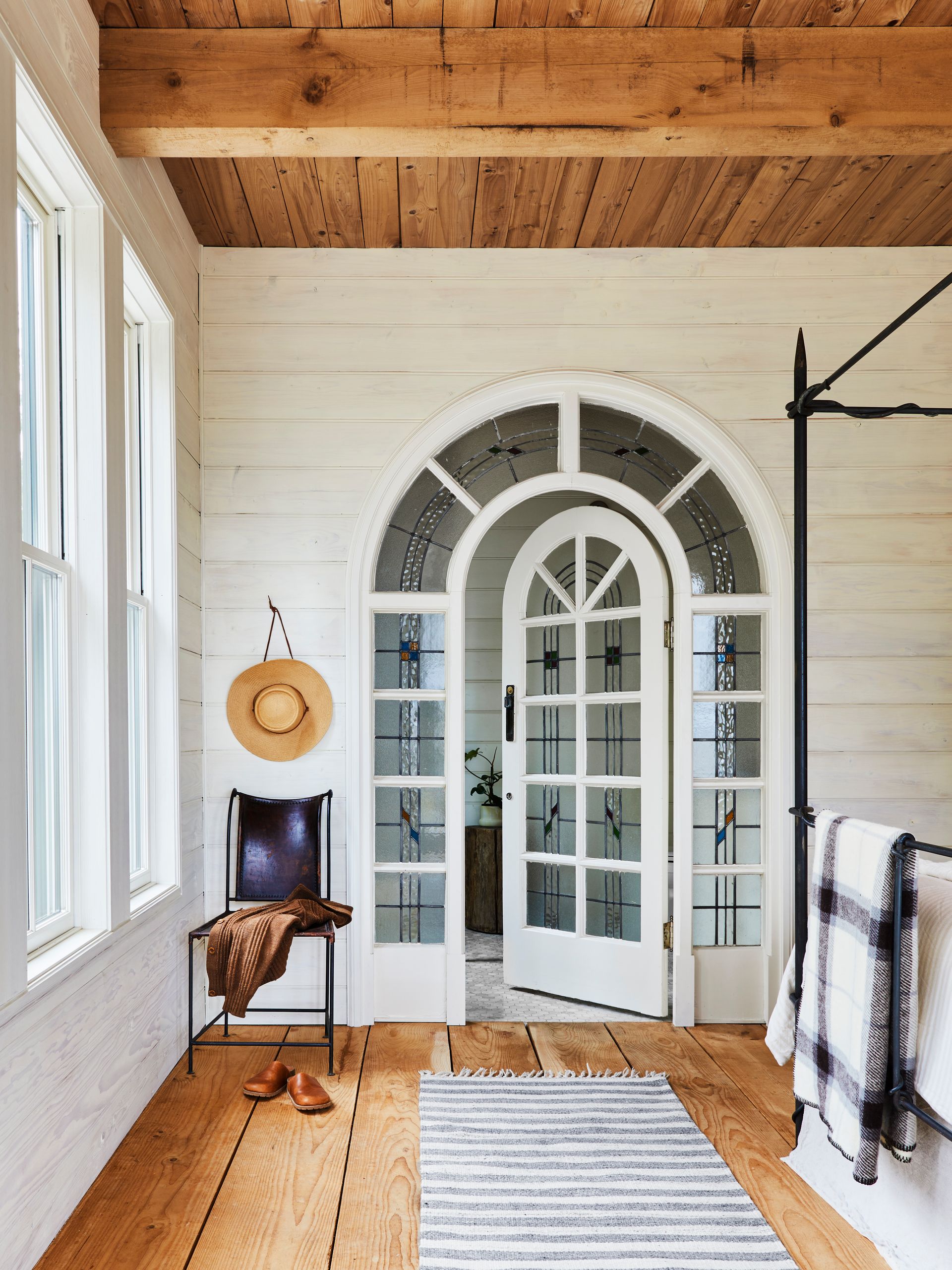 Bedroom with white-stained paneling on walls, wide plank wood flooring and ceiling, salvaged leaded glass arched door and wrought iron furnishings. 