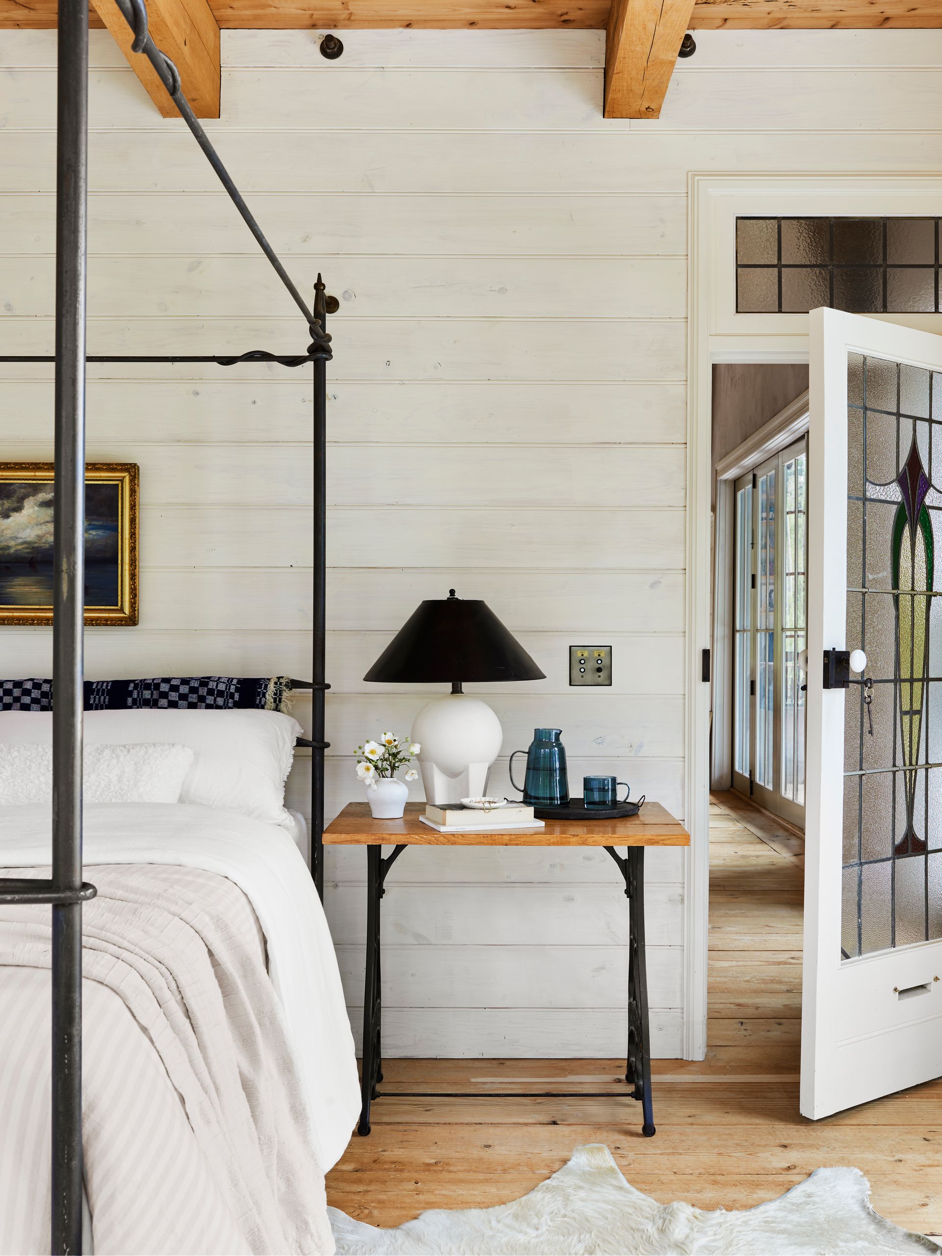 Bedroom with white-stained paneling on walls, wide plank wood flooring, salvaged leaded glass door and wrought iron furnishings. 
