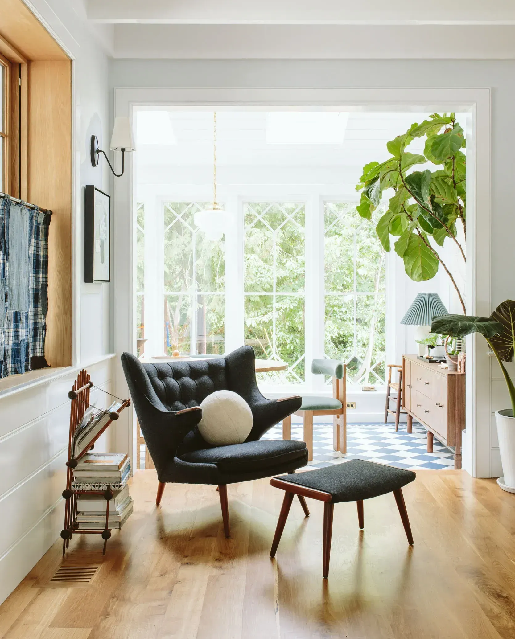 Cozy living room with a dark armchair, footstool, and a large plant near a bright sunroom.