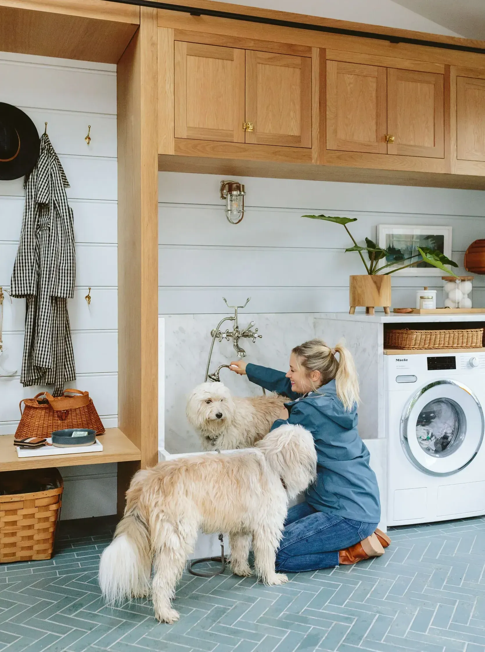 Woman bathing two fluffy dogs in a utility room with blue tile floor and wooden cabinetry.