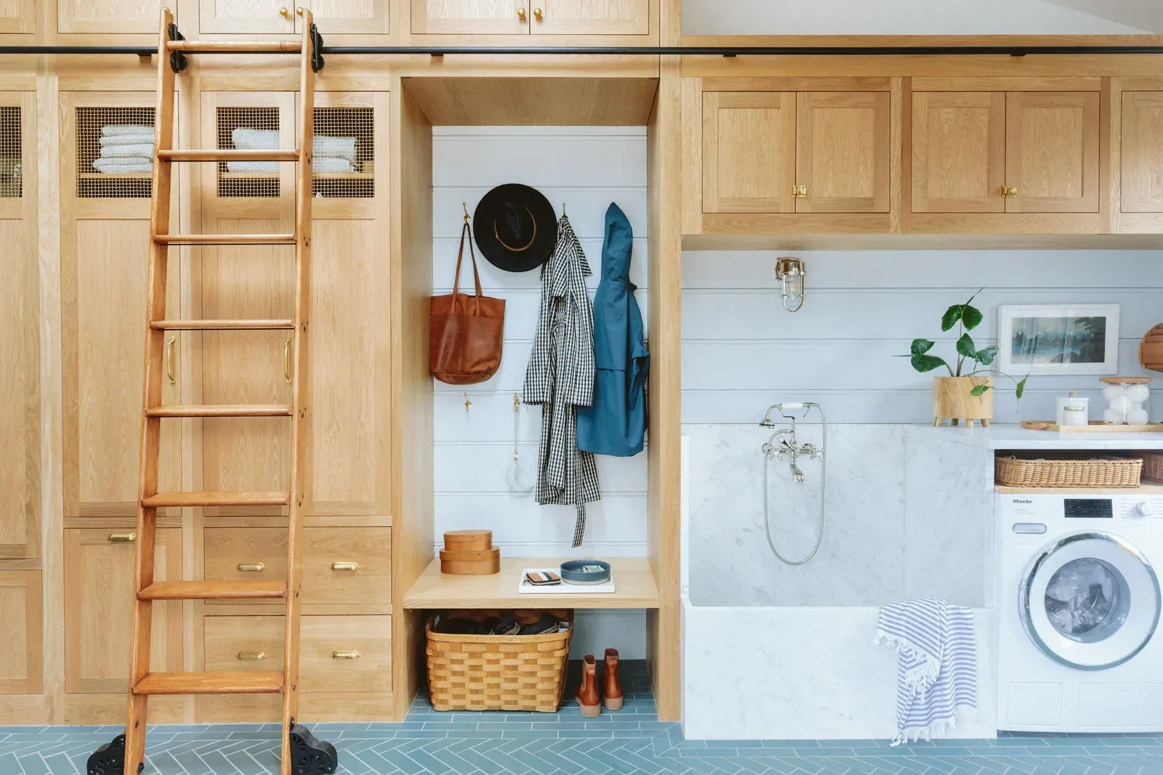 A laundry room with wooden cabinets, a bench, and a washing machine. Clothes and accessories hang on the wall.