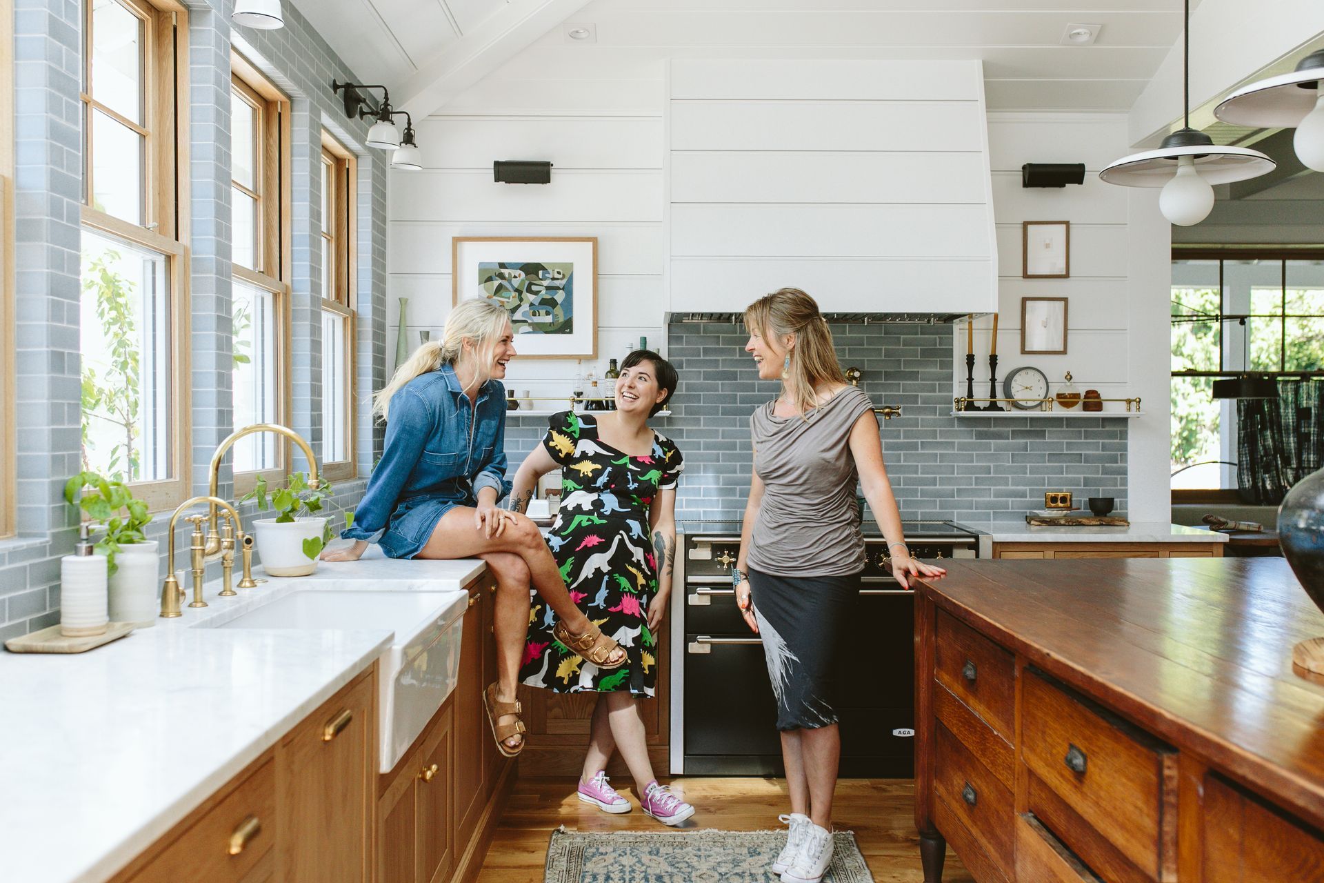 Three women laughing in a bright kitchen. One sits on the counter, two stand near the island.