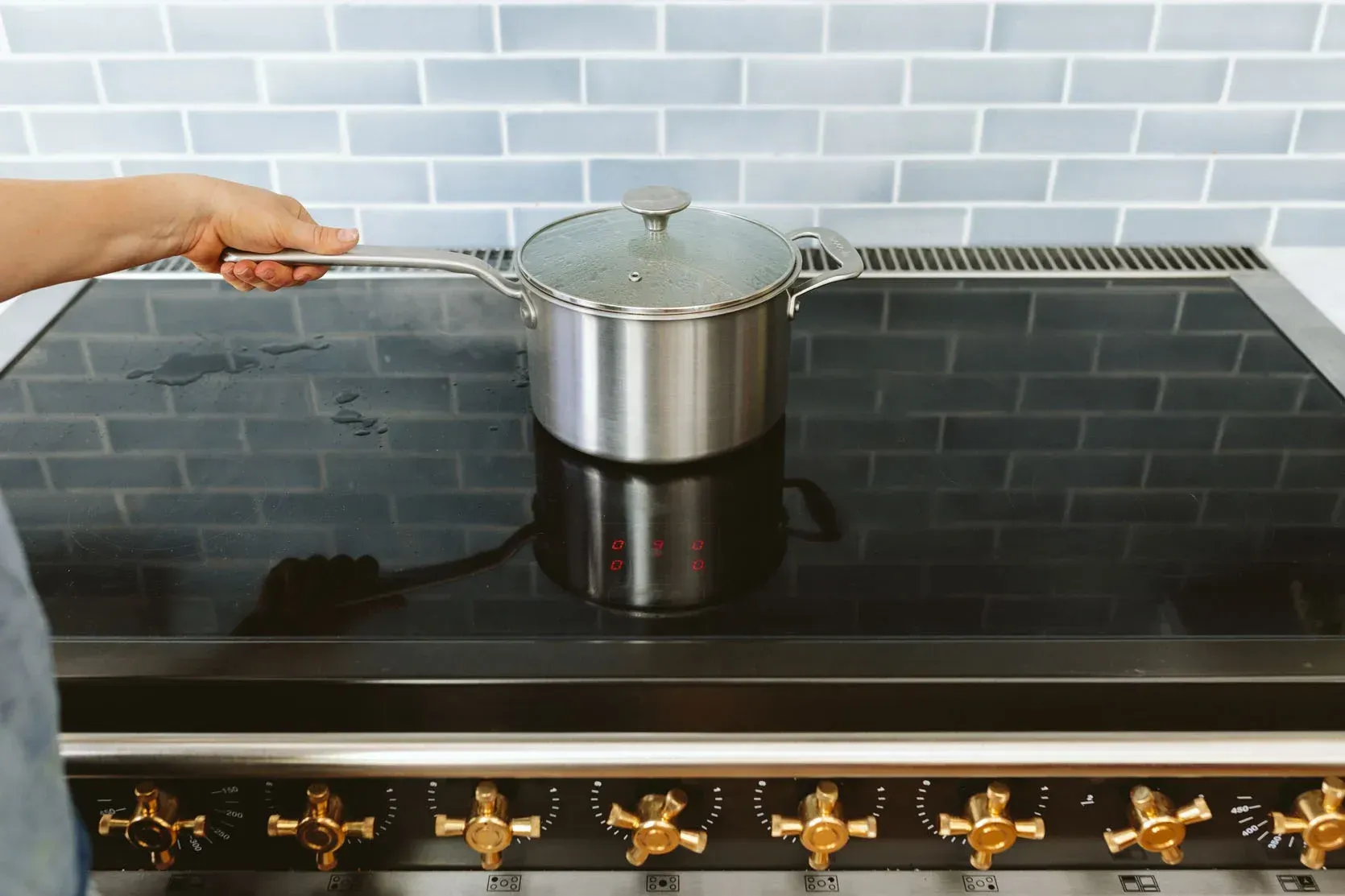 A person placing a stainless steel pot with a glass lid on a black stovetop.