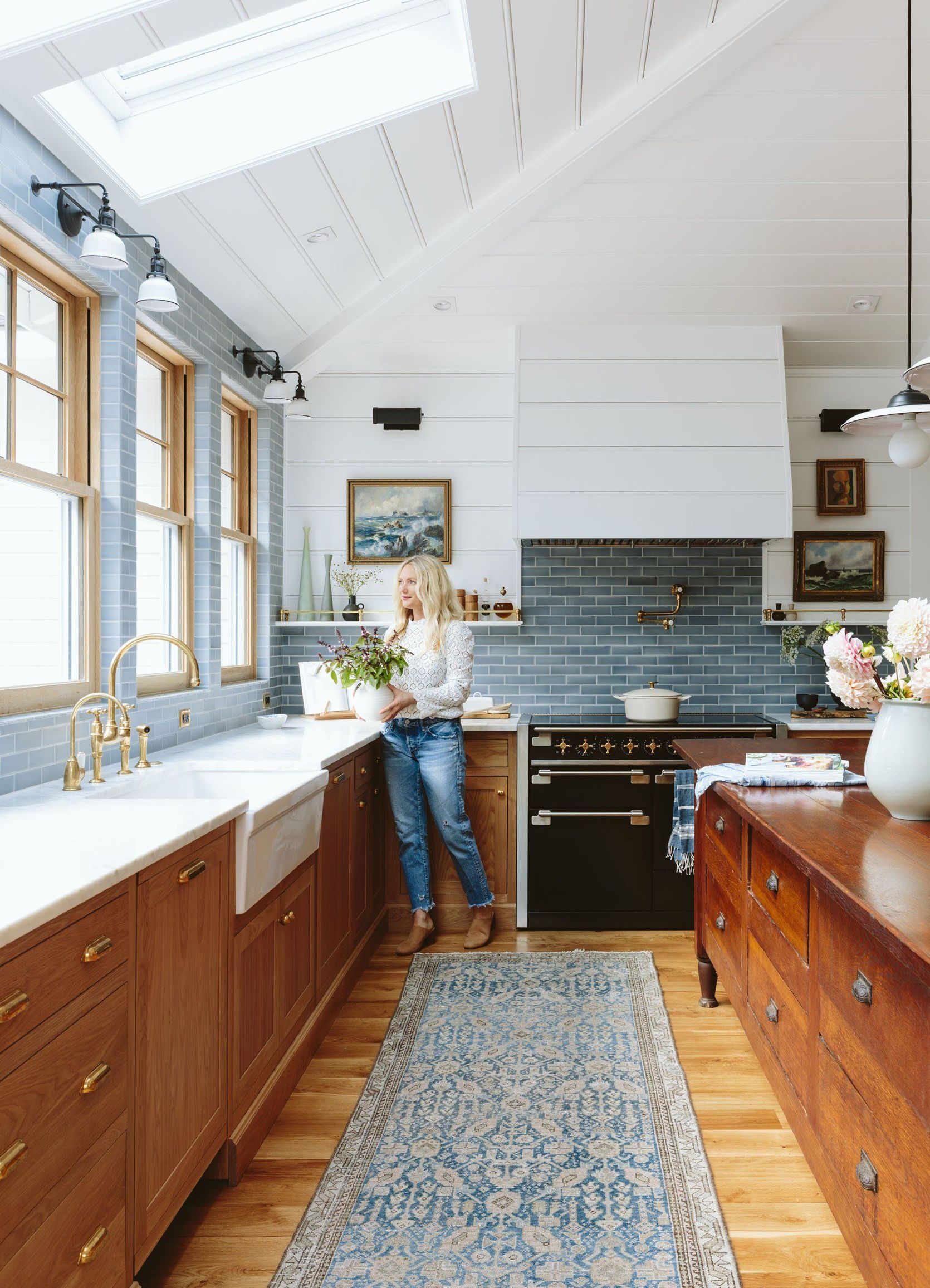 Woman in a light-filled kitchen stands by sink, light wood cabinets, blue tiled backsplash, vintage rug.