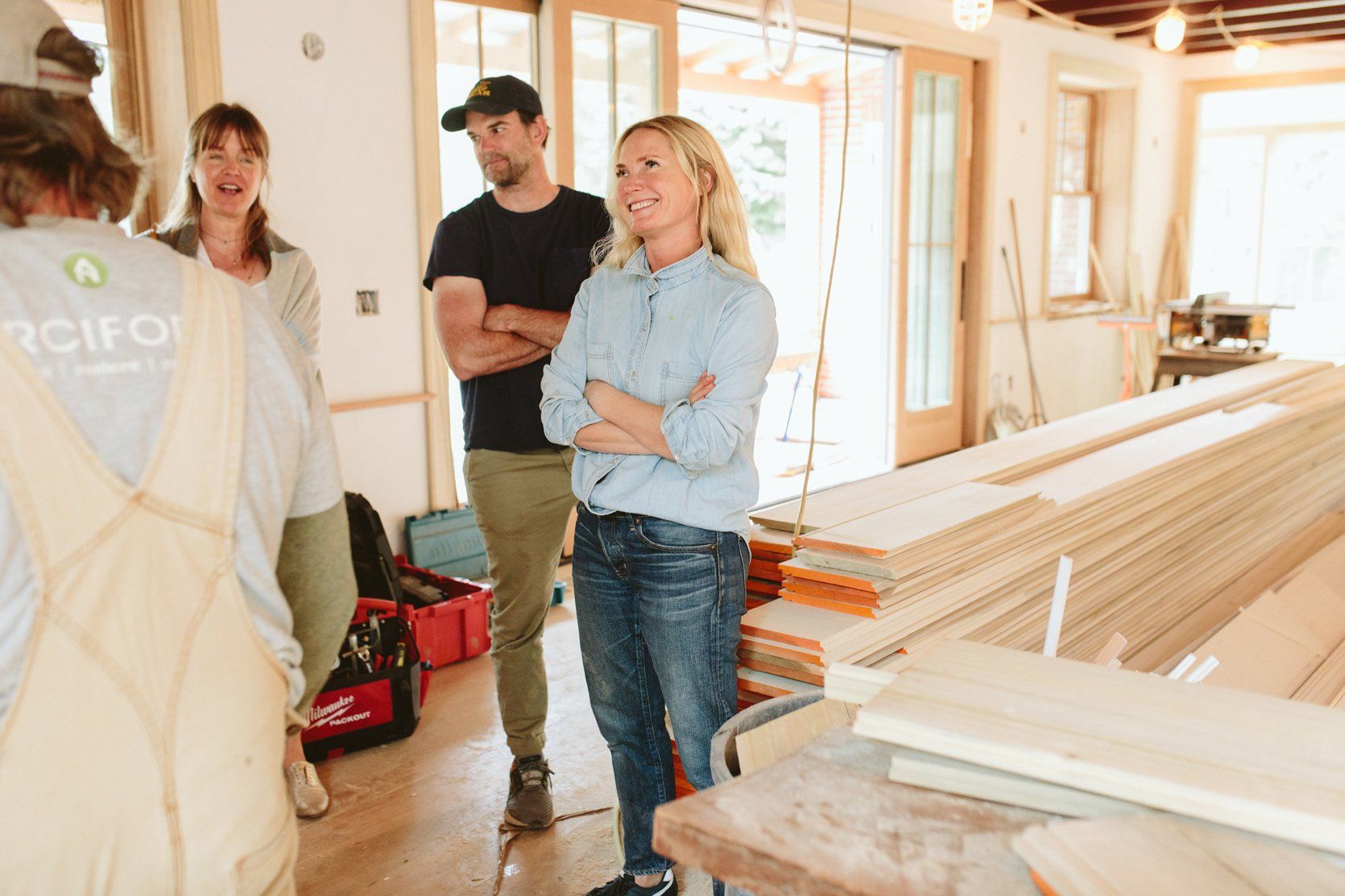 People in a home under construction, discussing plans. Woman in blue shirt smiles, arms crossed. Wood and tools visible.