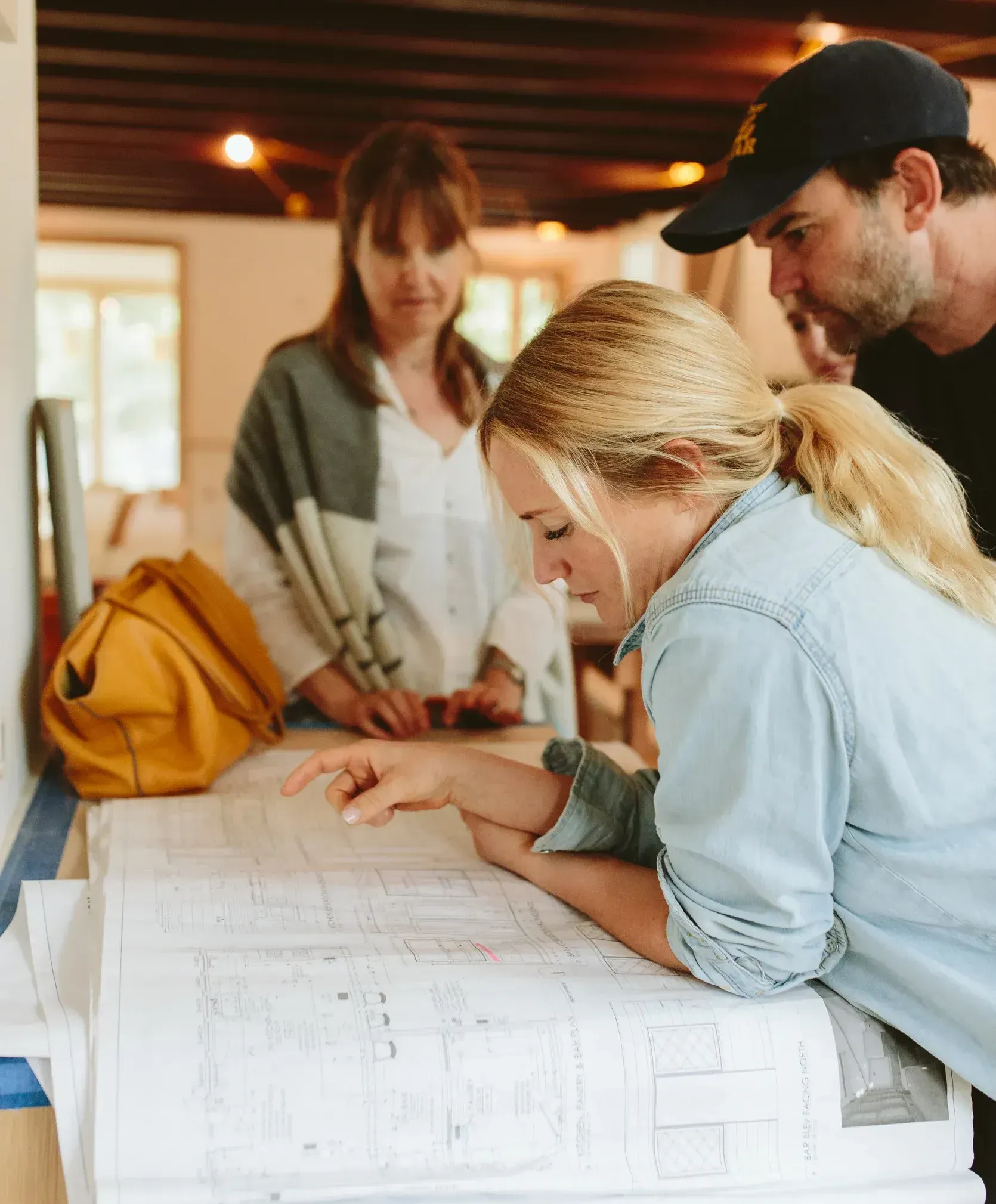 Three people reviewing blueprints at a construction site. Woman in denim shirt points, others observe.