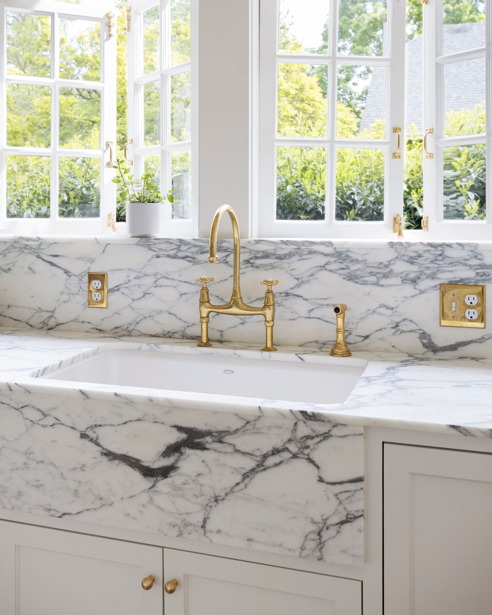 A close up of beautifully veined Arabescato marble at the kitchen sink, under a pair of windows that open to the garden. 