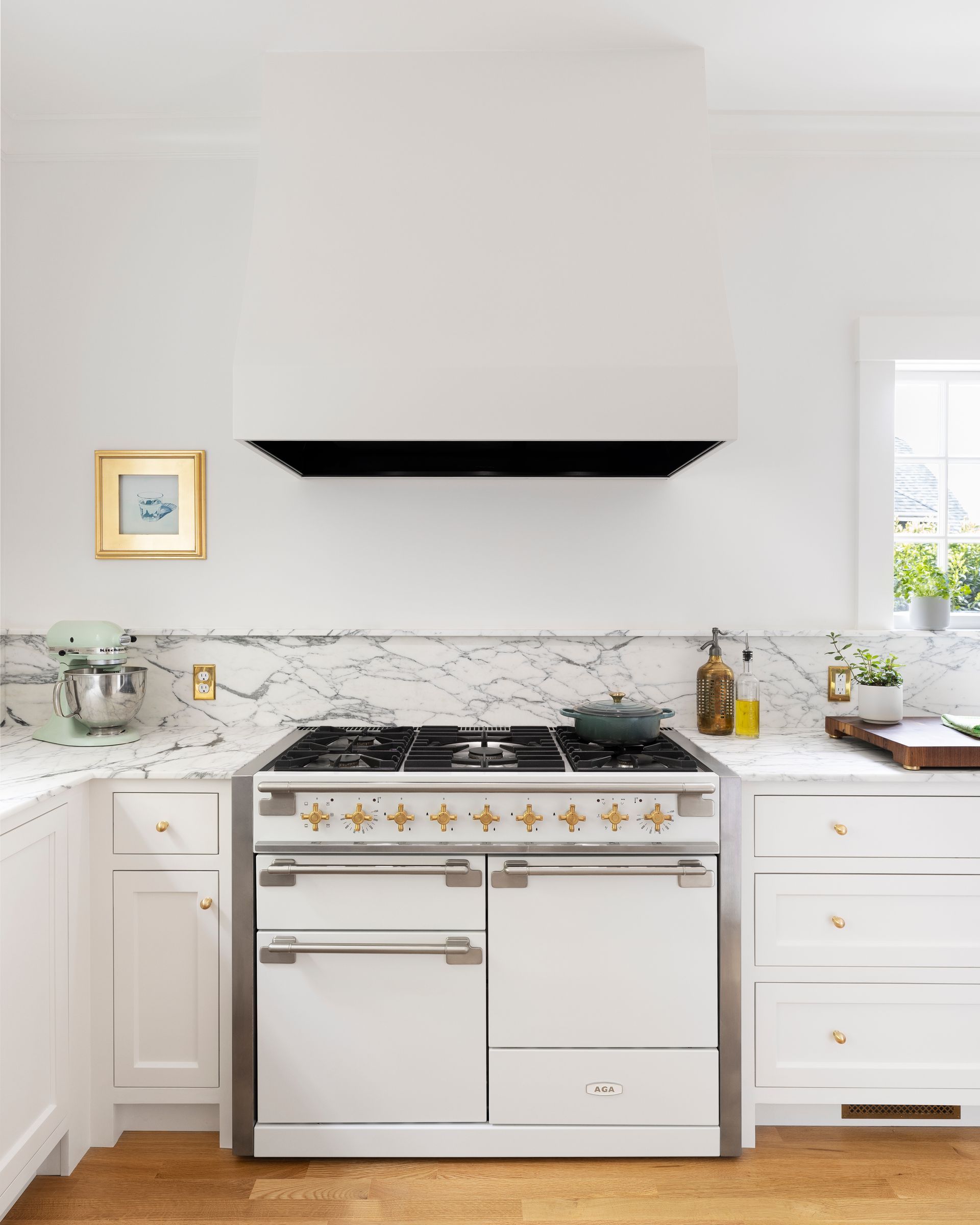 The range is a white, dual fuel Aga with brass knobs, with a Vent-A-Hood range hood with a custom cover to blend into the wall. 