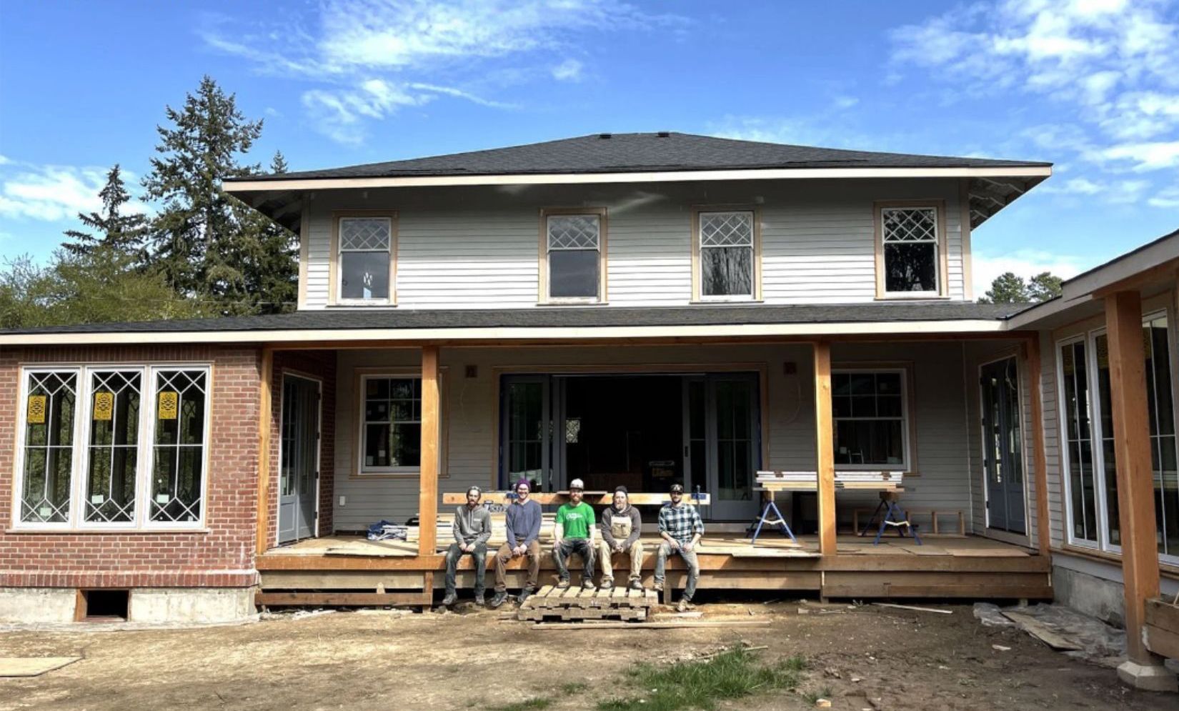 Construction workers posing on a porch, wooden structure being added to a white house. Sunny day.