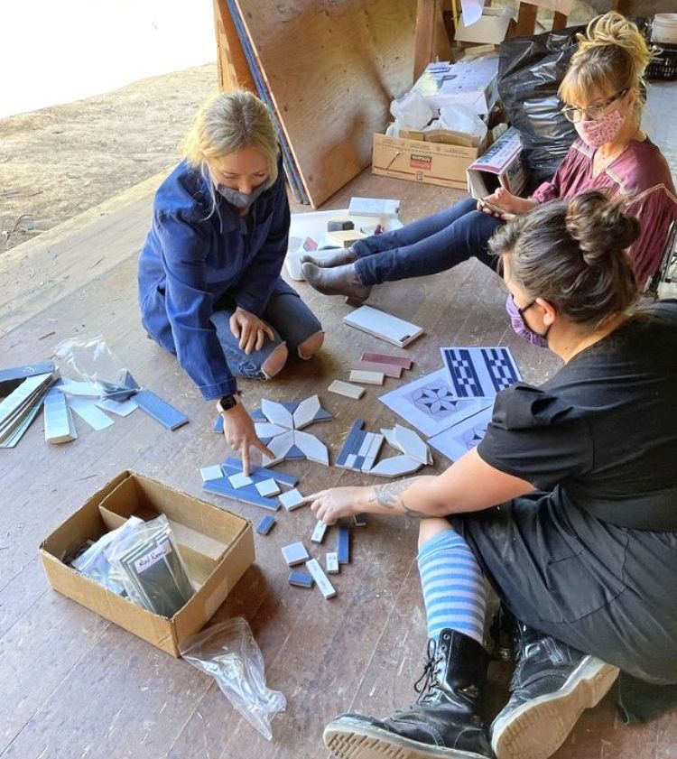 Three women wearing masks plan a tile mosaic on a floor. One points at tiles, others seated nearby.