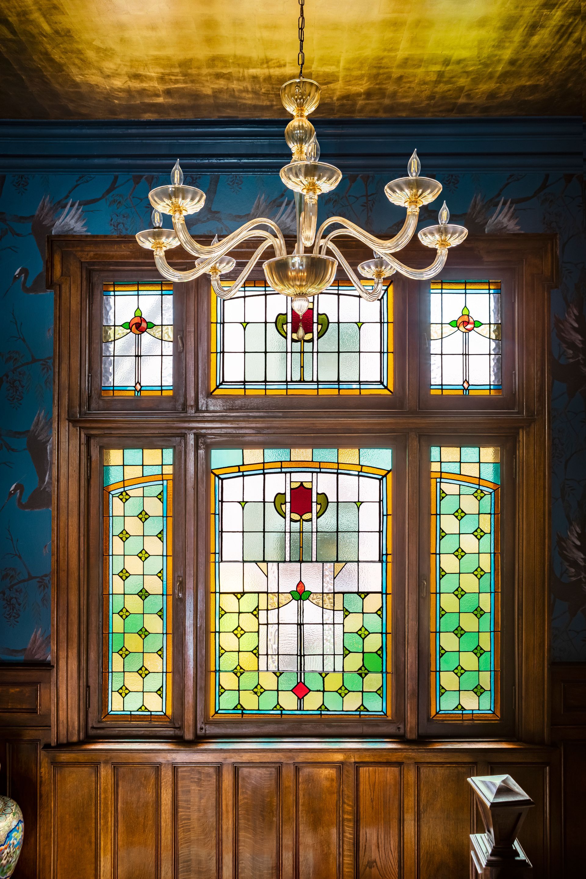 Large stained glass windows surrounded by oak millwork behind a Murano glass chandelier, all under a brass-leafed ceiling.