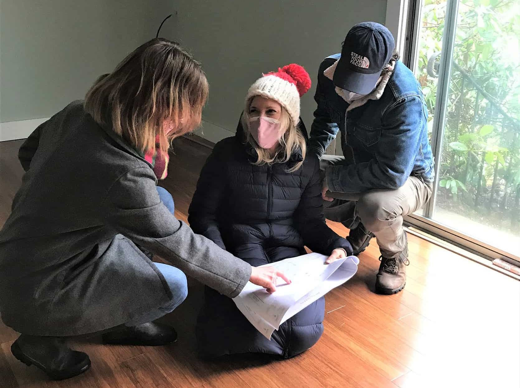 Three people reviewing blueprints in a room with wood floors and a large window. All are wearing masks and winter clothing.