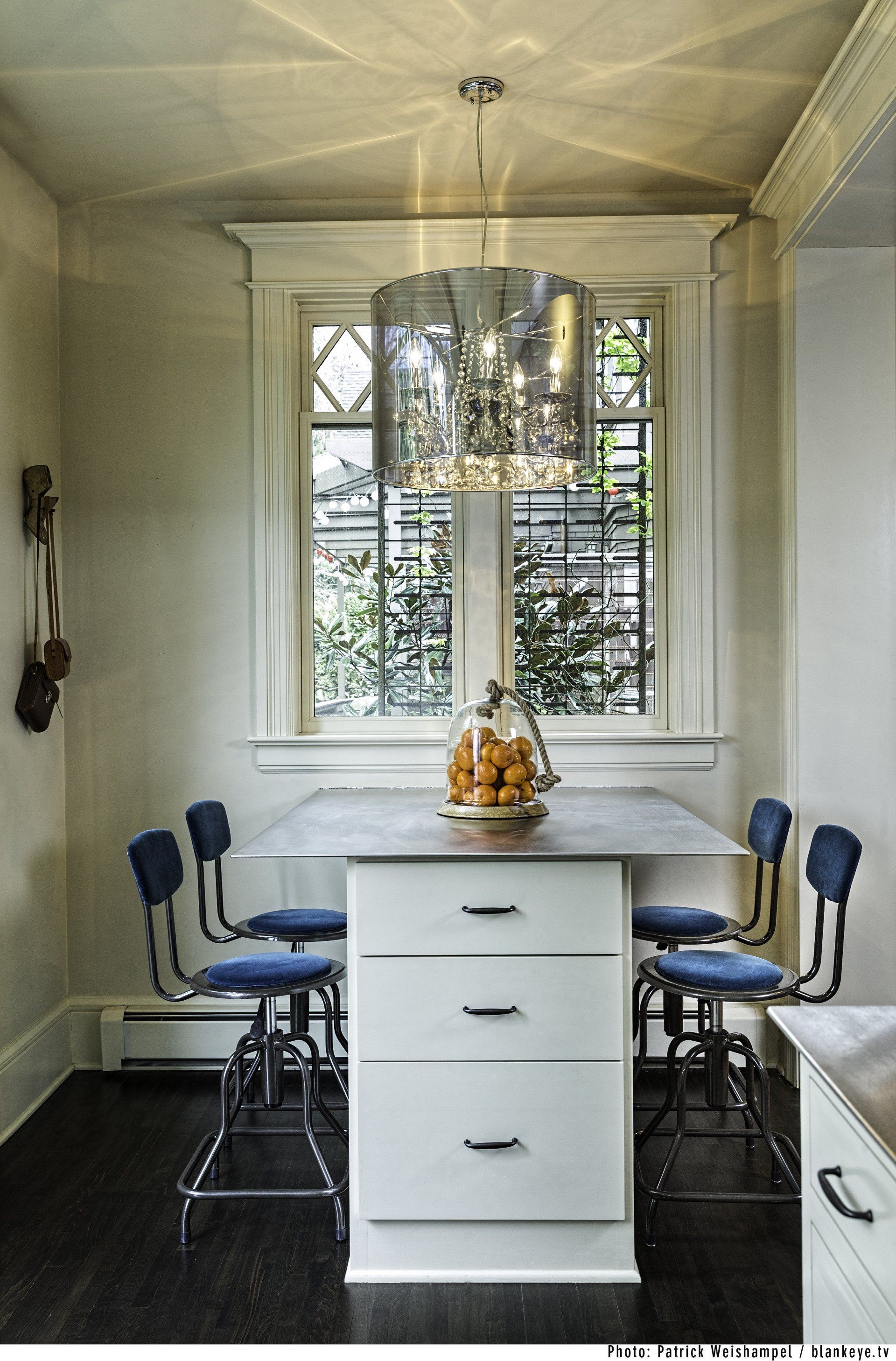 View from the kitchen of the dining nook, with a tall custom island with extra-deep drawers and upholstered swivel stools under a large window and glistening drum pendant.