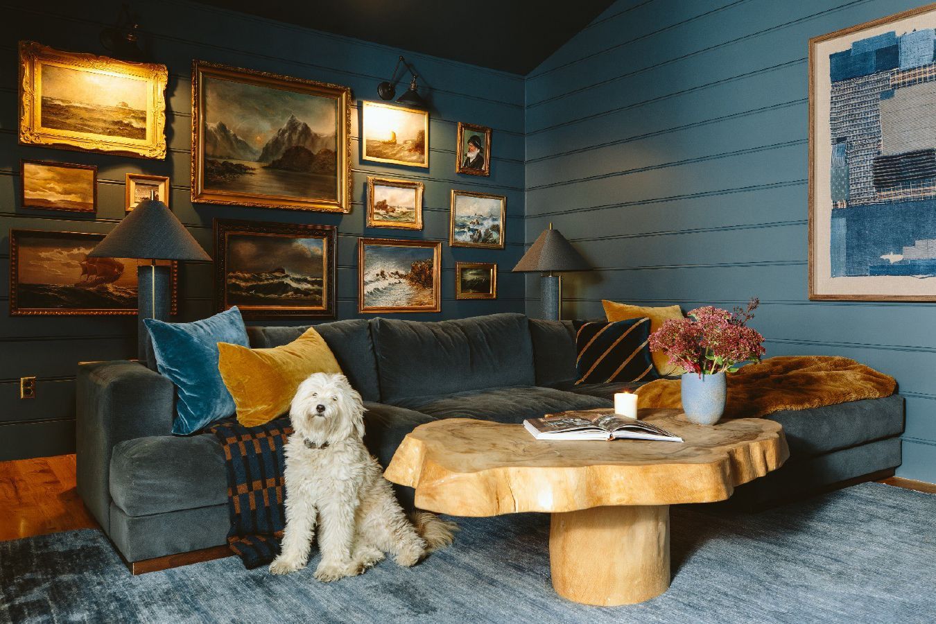 Cozy living room with blue walls, framed landscape art, a gray couch, a wooden table, and a fluffy dog.