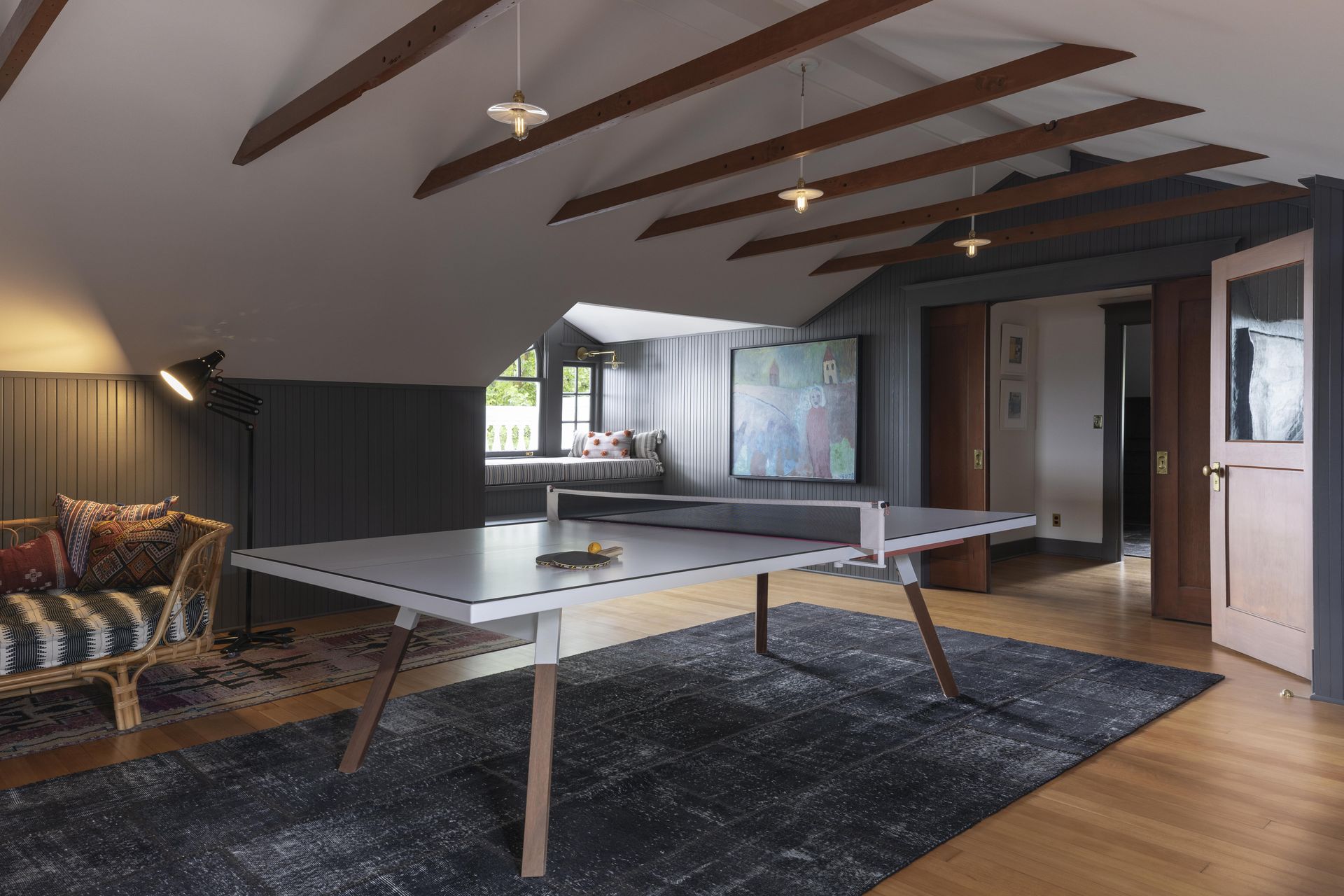 Remodeled attic with exposed collar ties, beadboard paneling, wood floors, a ping pong table, and casual furnishings with a window seat beyond.