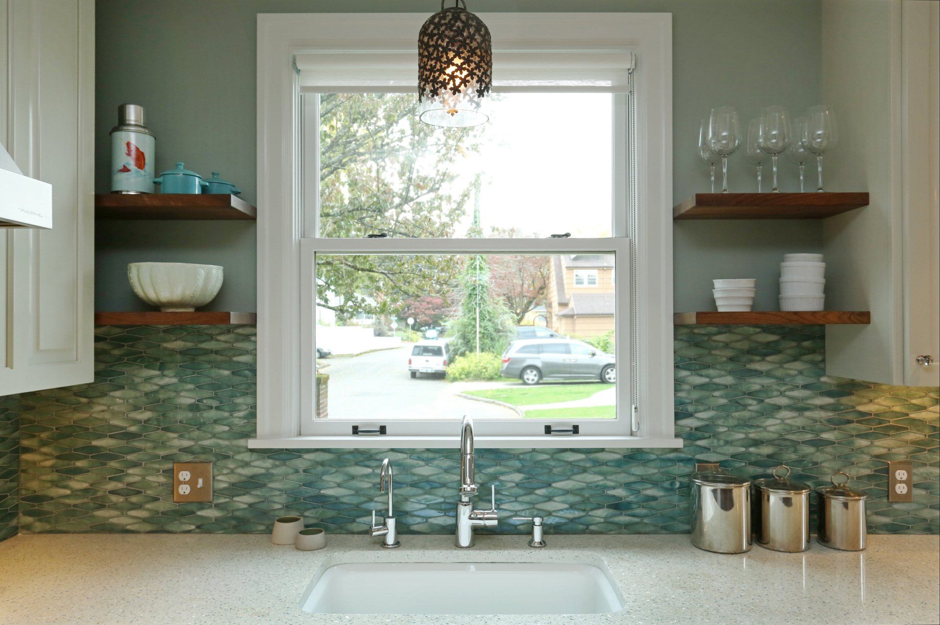 Looking out the kitchen sink window, which is flanked by shelving with sea-hued glass backsplash tile.