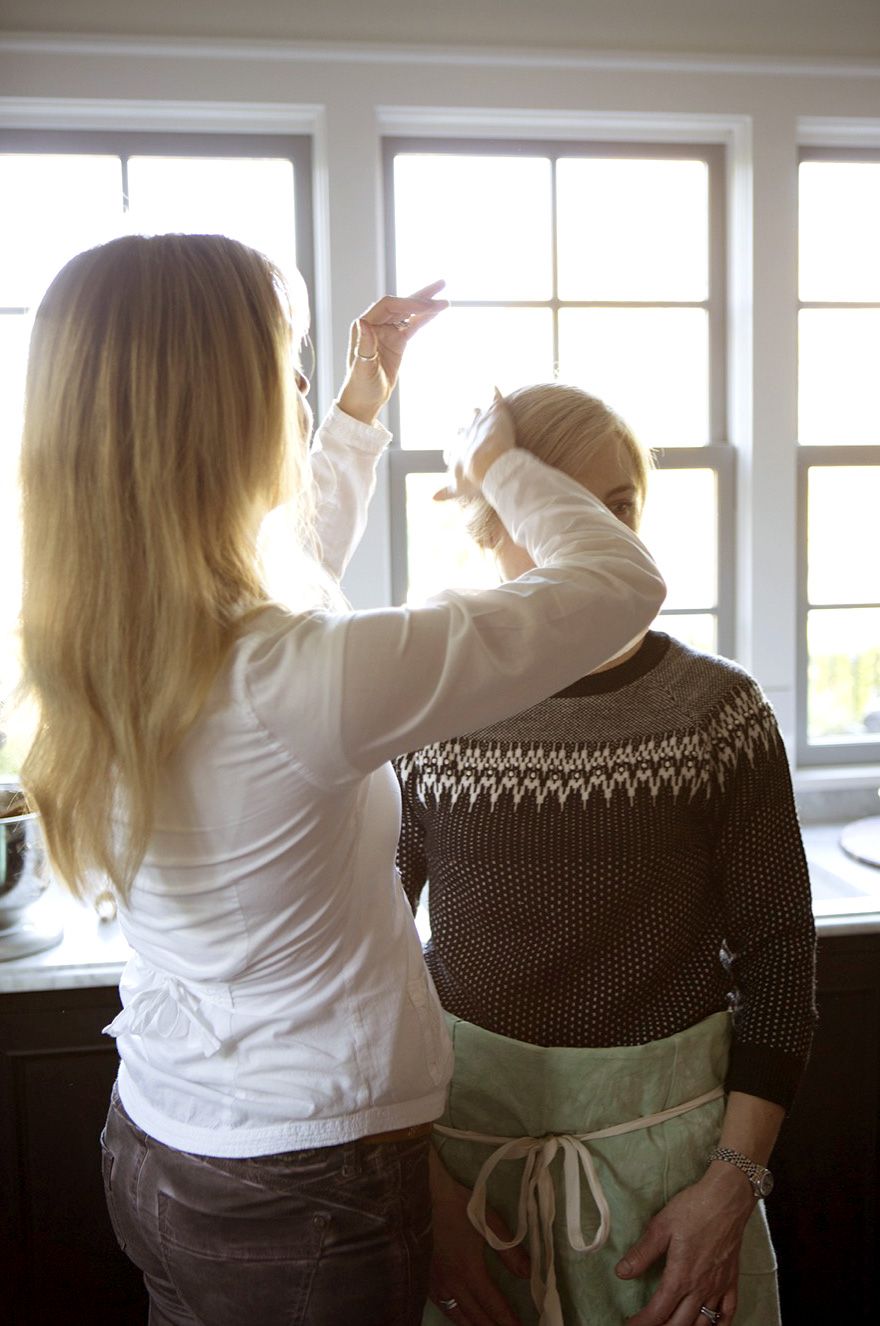Woman styling another person's hair in a bright kitchen by a window