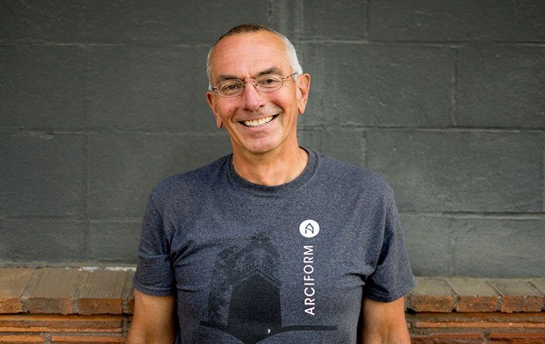 A man wearing a gray t-shirt and glasses is smiling in front of a brick wall.