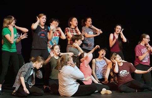 A group of young people are sitting on the floor on a stage.