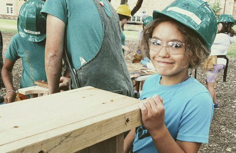A young girl wearing a hard hat and glasses is standing next to a wooden table.