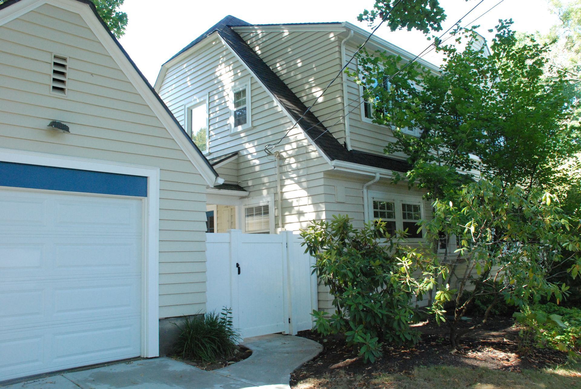 White suburban house with garage, white fence, and leafy bushes beside a driveway