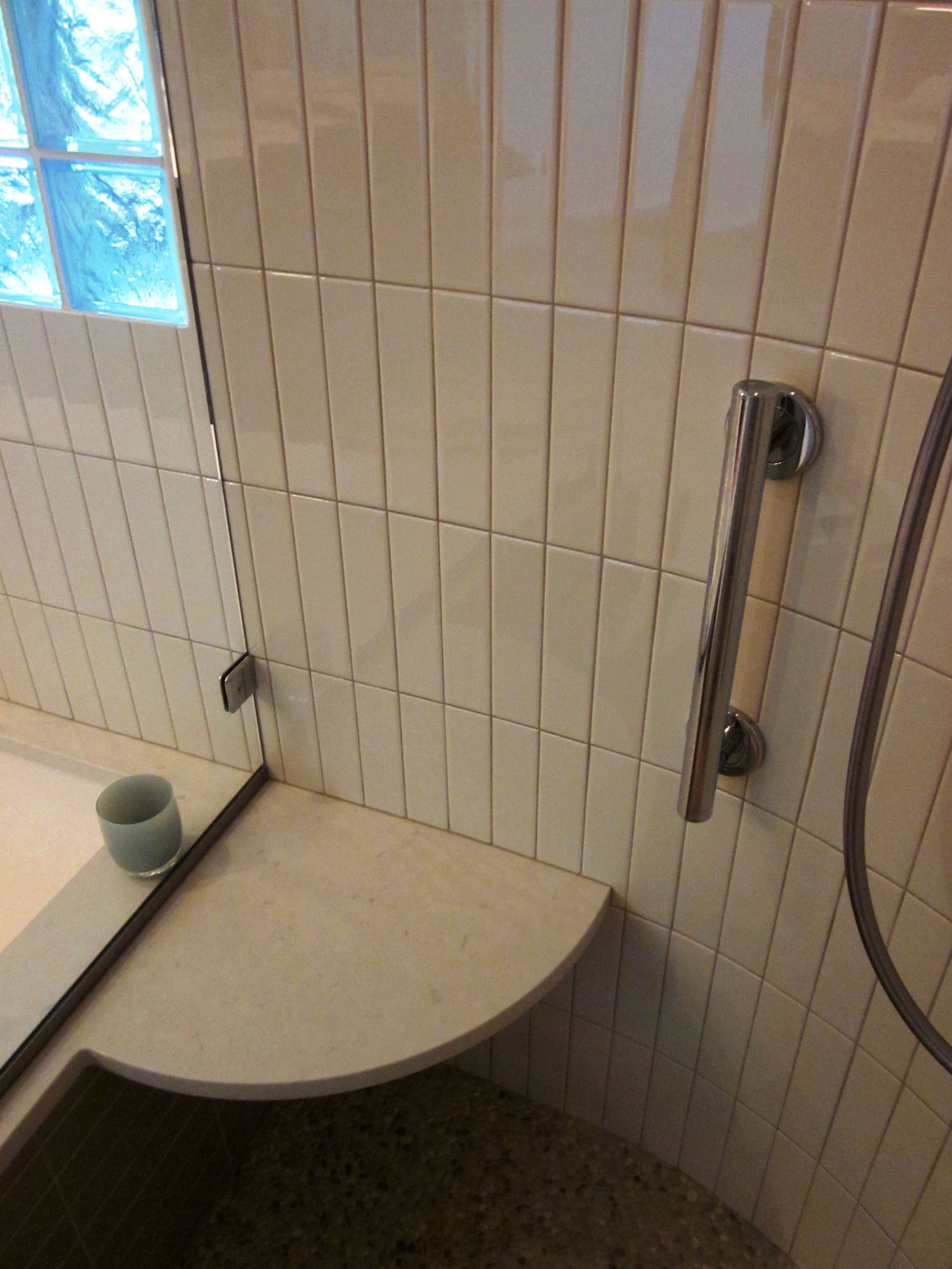 Small tiled shower stall with a curved bench and handheld showerhead beside a frosted window.