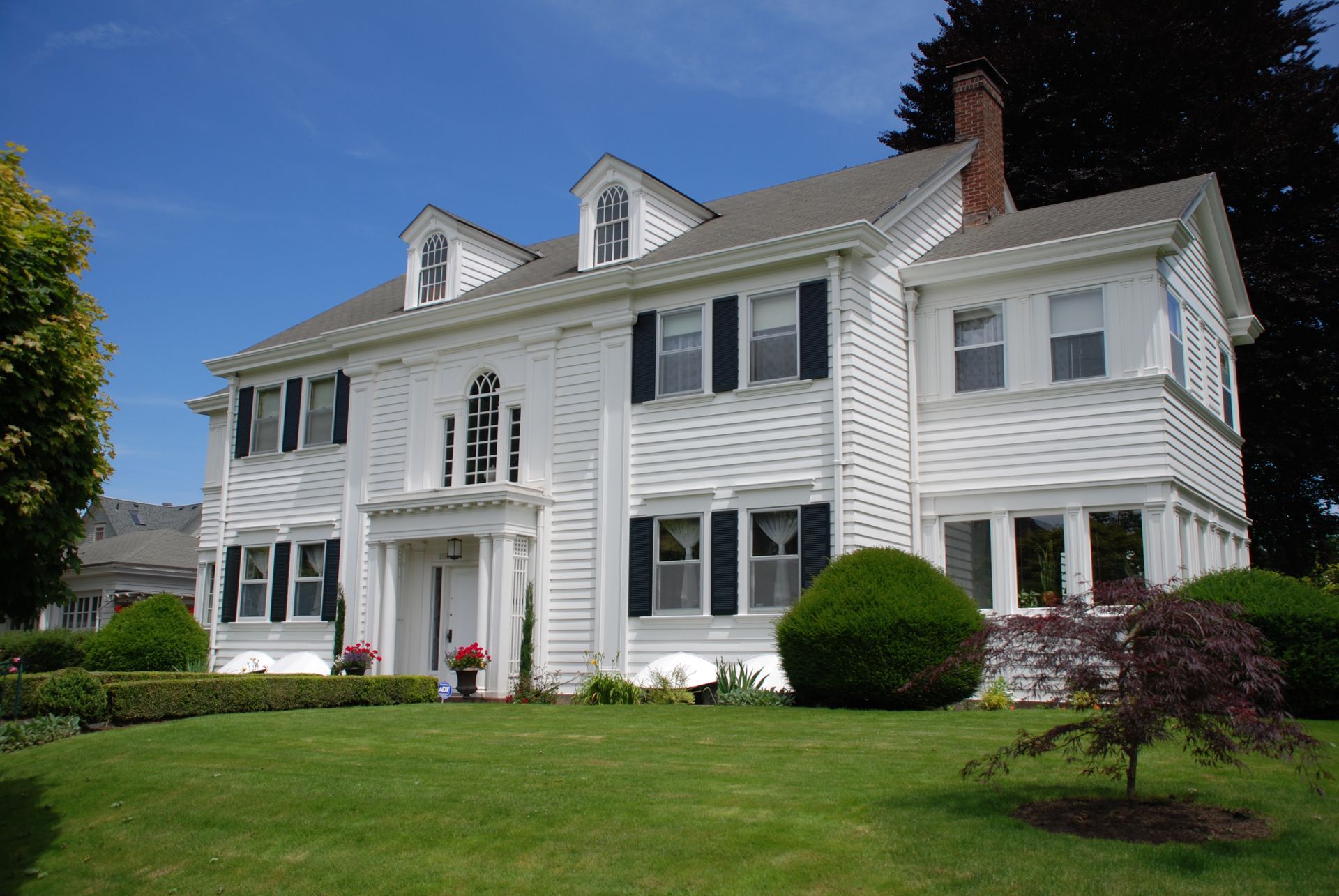 White two-story colonial house with shutters, dormer windows, and a manicured green lawn under a blue sky