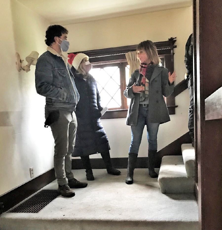Three people standing in a house entryway. One woman speaks, others listen. Staircase visible; Christmas decor.