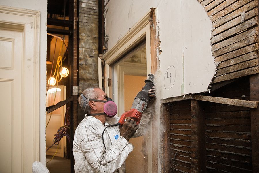 Worker in respirator using a power drill to remove brick from an interior wall.