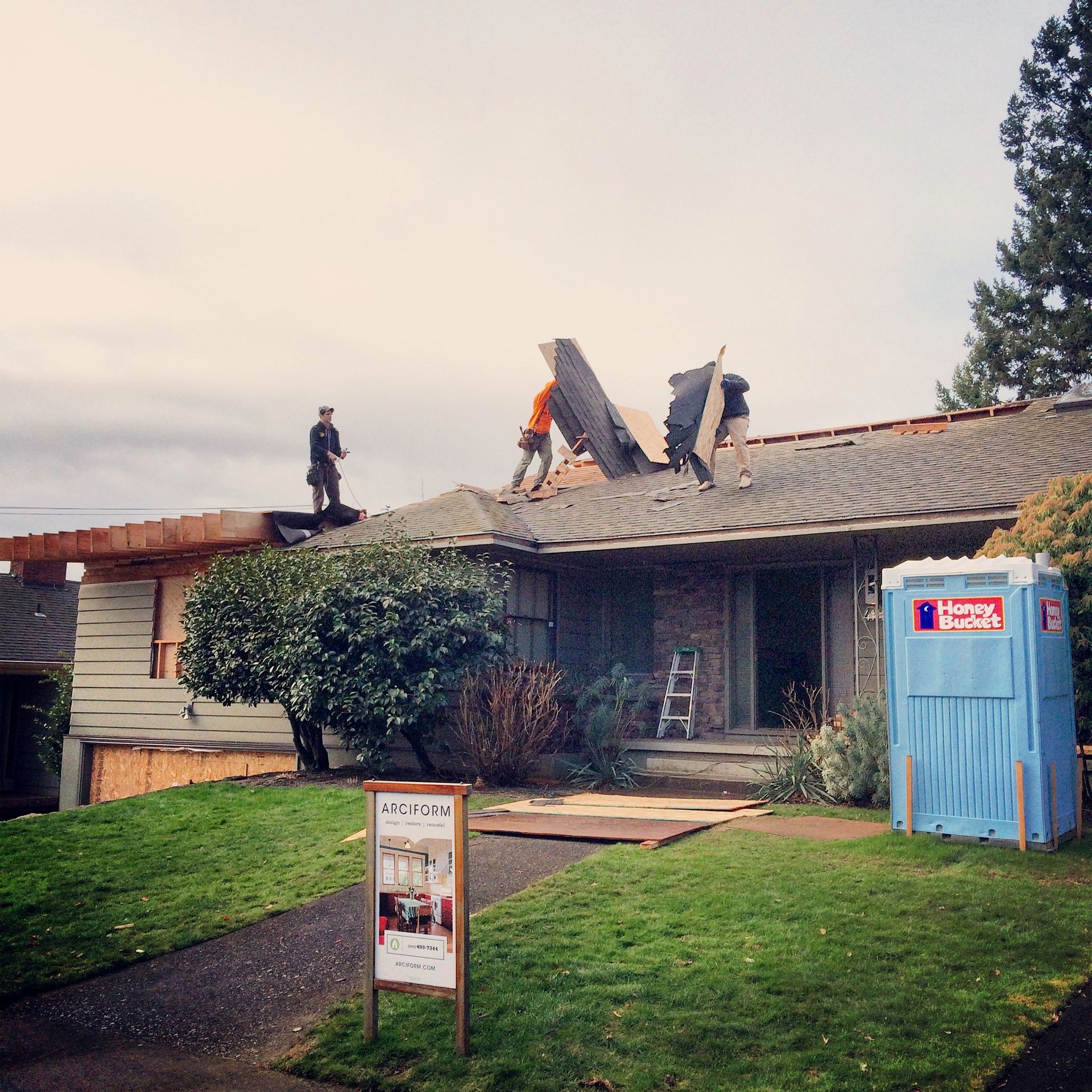 Workers on a roof repairing a low building beside a blue portable toilet.