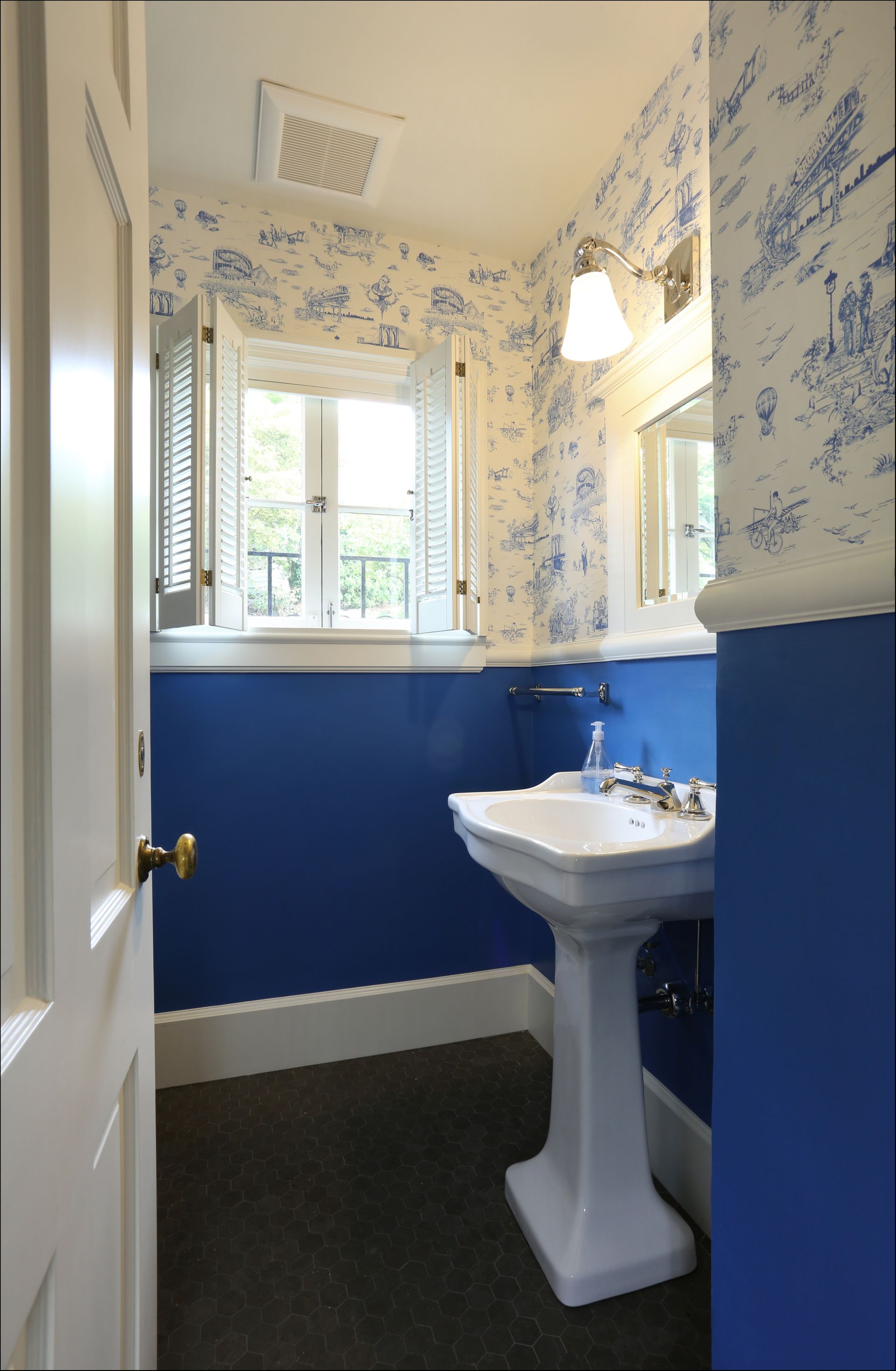 Small bathroom with blue wainscoting, floral wallpaper, pedestal sink, and windowed shower area.