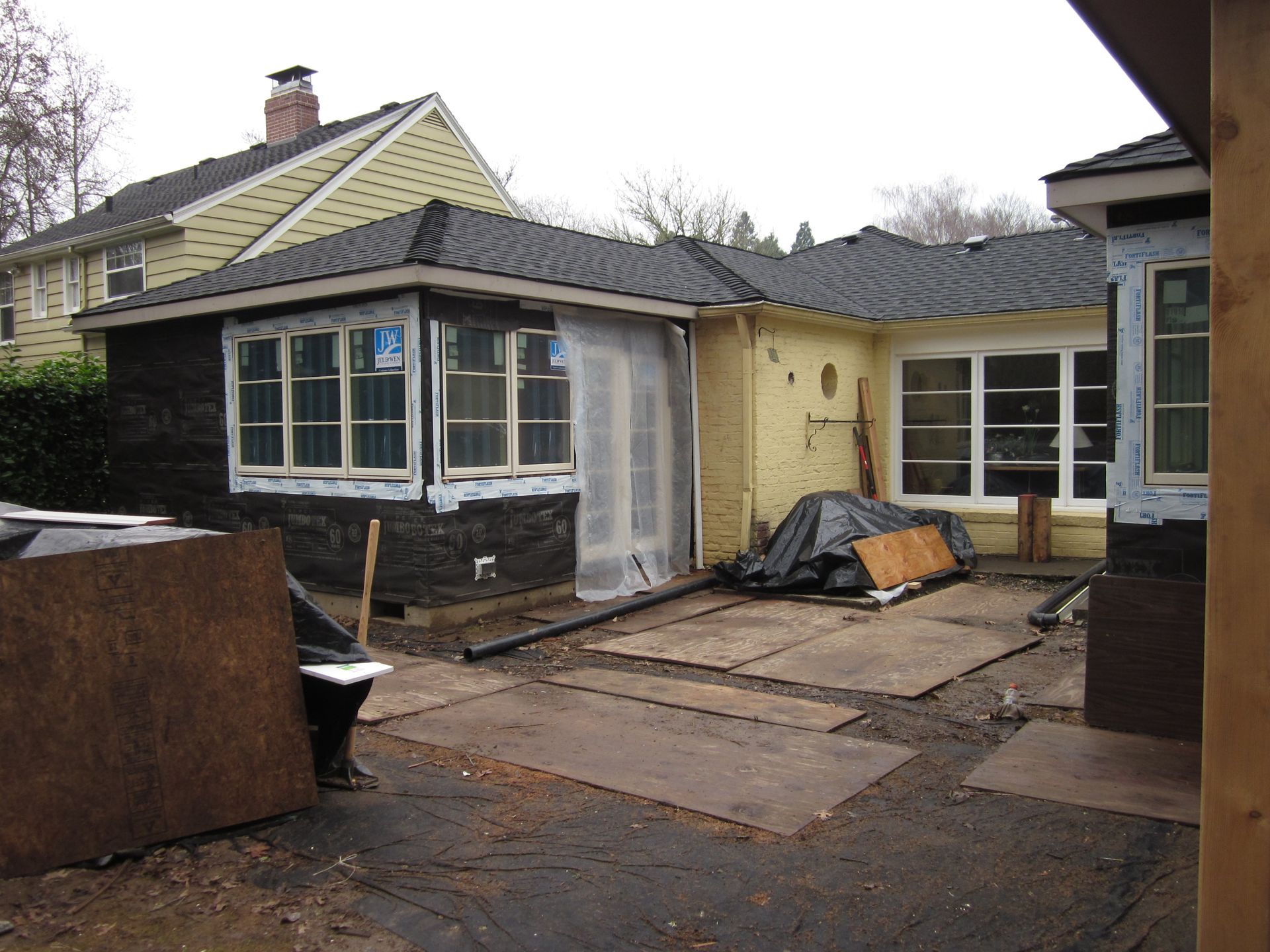 Partially renovated house exterior with roof work, boarded windows, and construction materials in the yard
