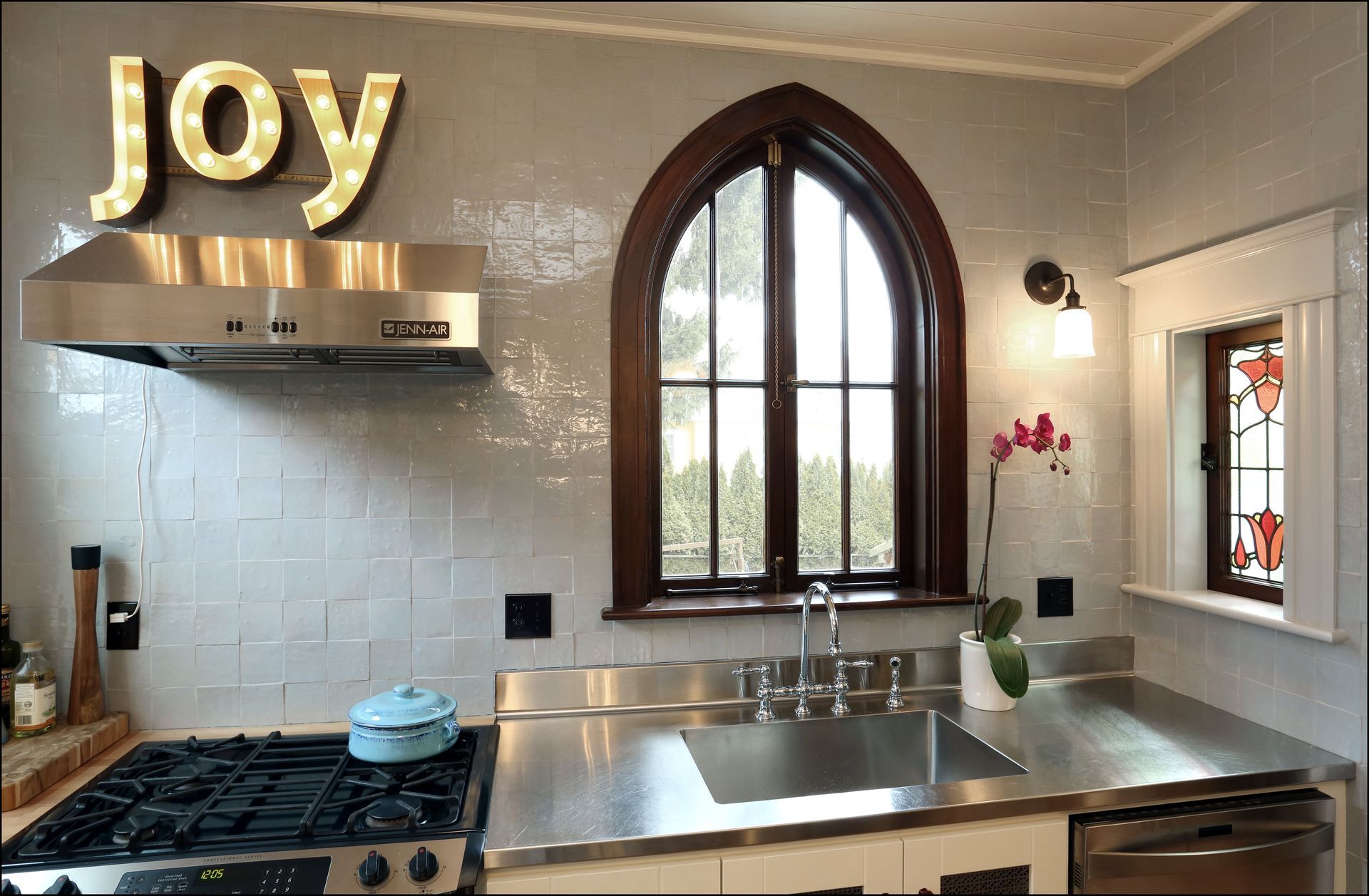 Bright modern kitchen with stainless steel appliances, arched window, and gold “joy” sign on the wall