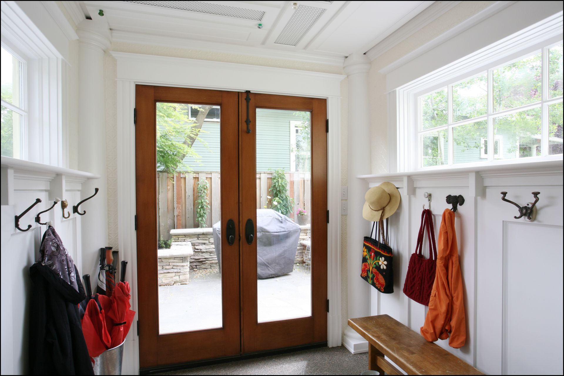 Bright mudroom with wooden double doors, white walls, hooks, bench, coats, and a glimpse of patio outside.
