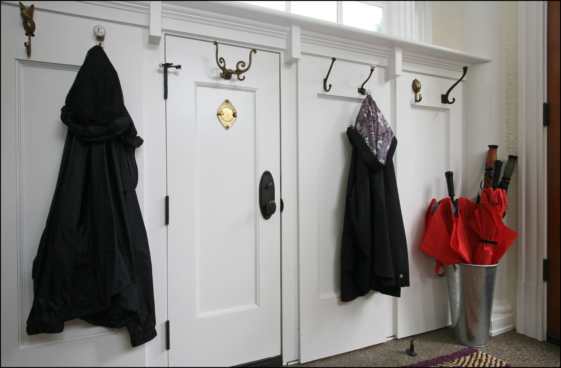 Black coats and red umbrellas hang on hooks in a white entryway with cabinet doors and tile floor.