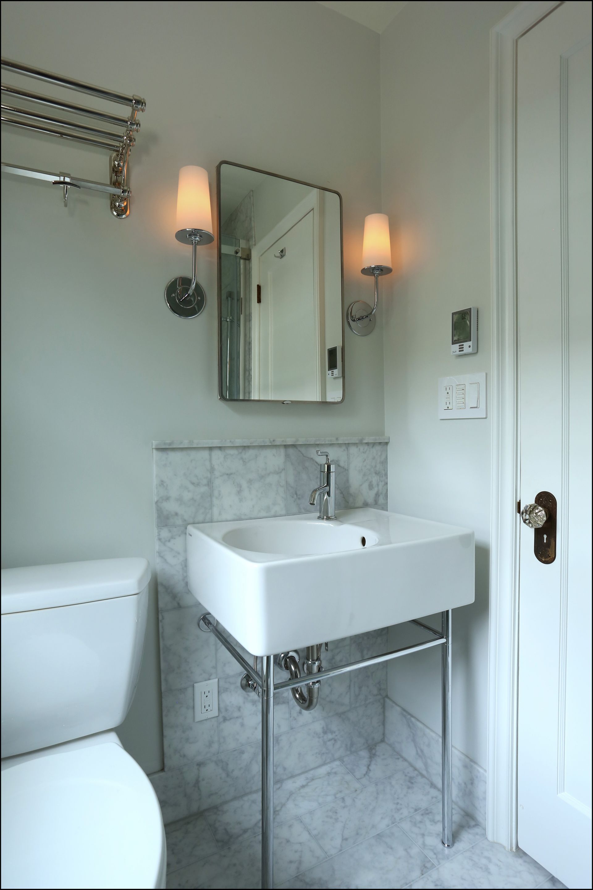Small bathroom with pedestal sink, mirror, sconces, marble tile, and toilet visible on the left.