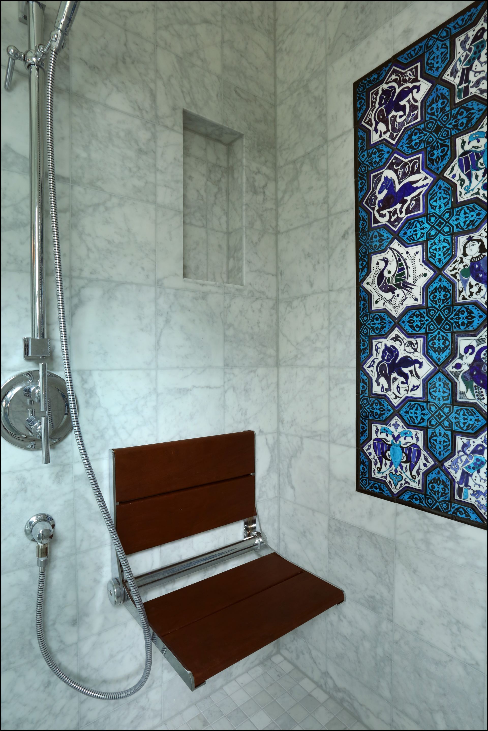 Shower with handheld sprayer, tiled walls, wooden fold-down seat, and decorative blue patterned tile panel