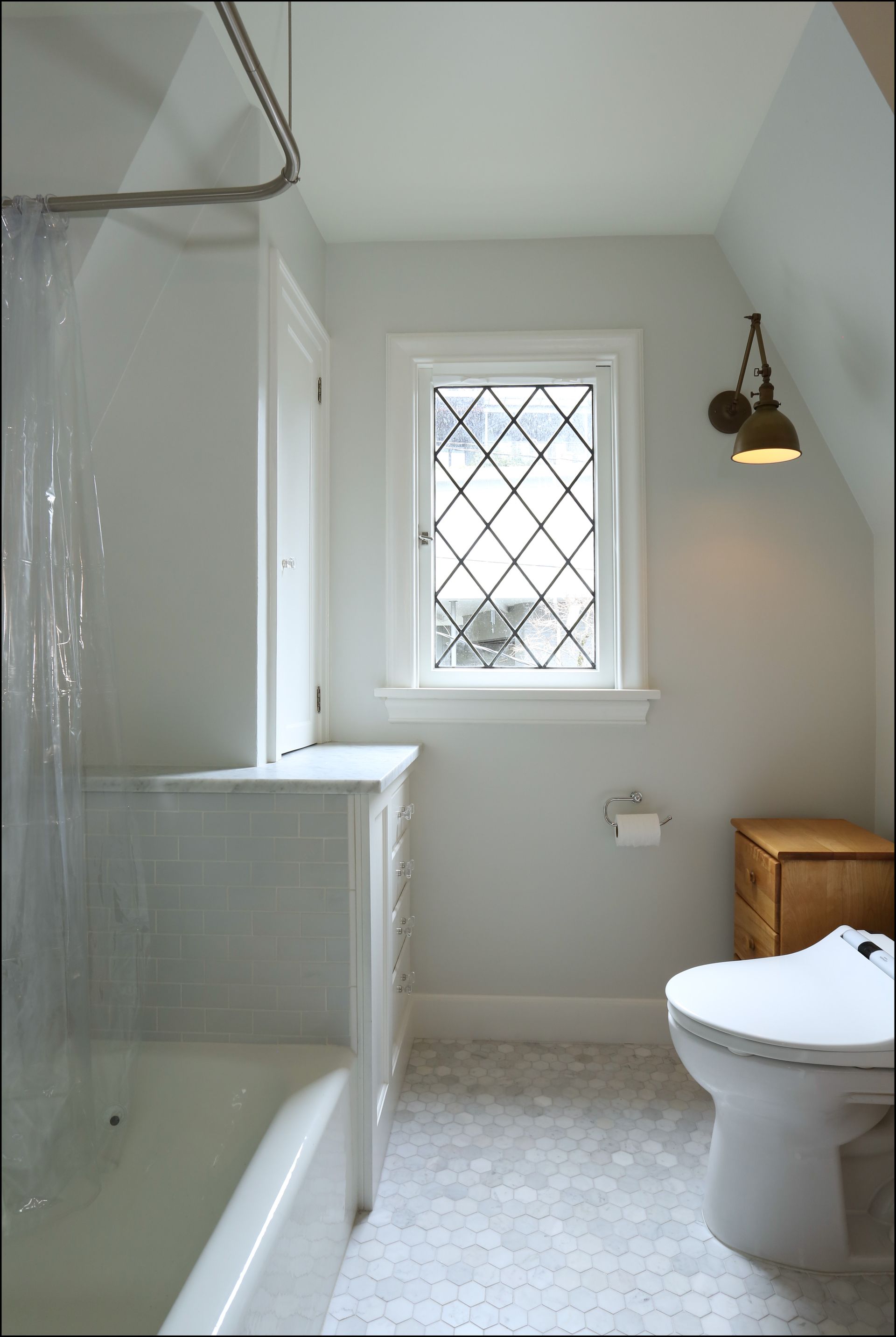Small white bathroom with a bathtub, toilet, window, and wall light.