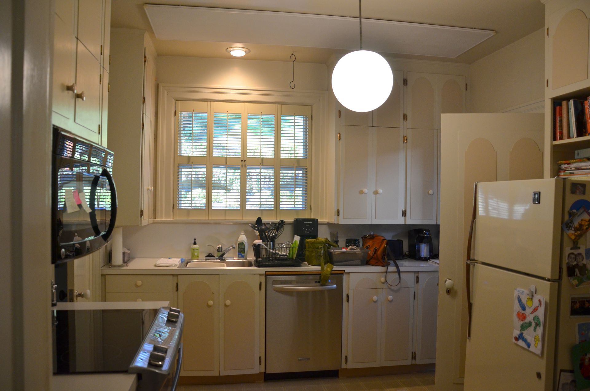 Bright kitchen with white cabinets, a central window, hanging globe light, and a refrigerator on the right.