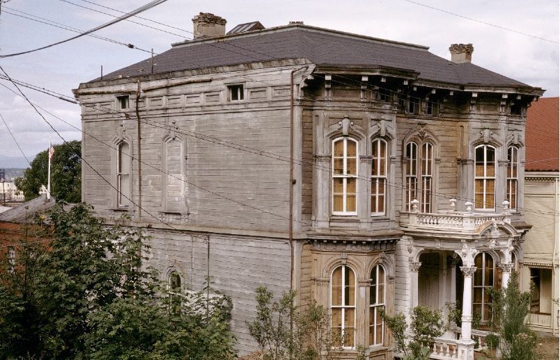 Old weathered gray two-story house with ornate windows, shown from the front-left on a quiet street.