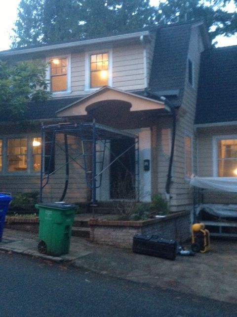 Two-story house with a front porch scaffold, green trash bins, and warm lights at dusk