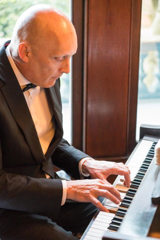 A man in a tuxedo is playing a piano in front of a window.