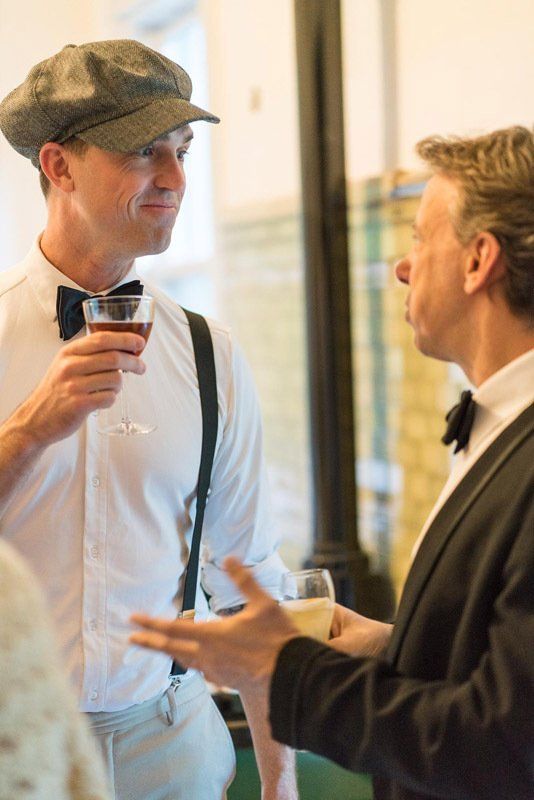 A man in a hat is holding a glass of wine while talking to another man in a tuxedo.