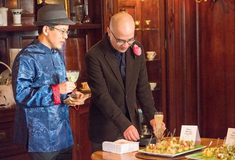 Two men are standing next to each other looking at food on a table.
