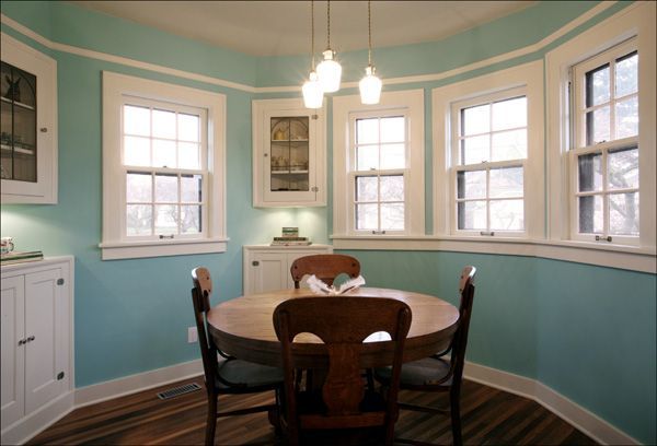 Bright dining room with teal walls, round table, wooden chairs, and large windows with white trim