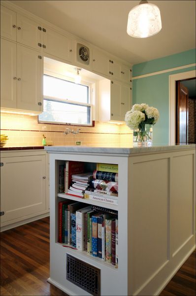 Bright white kitchen with a central island, bookshelves, and a vase of white flowers by a window.