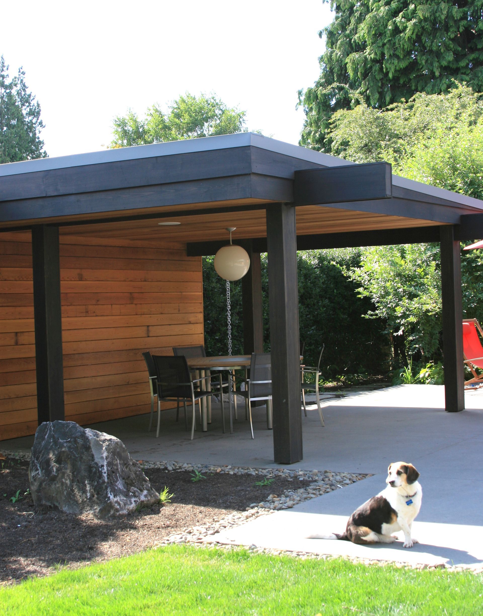 Patio with black-roofed pergola, outdoor table and chairs, and a small dog sitting on the concrete path.