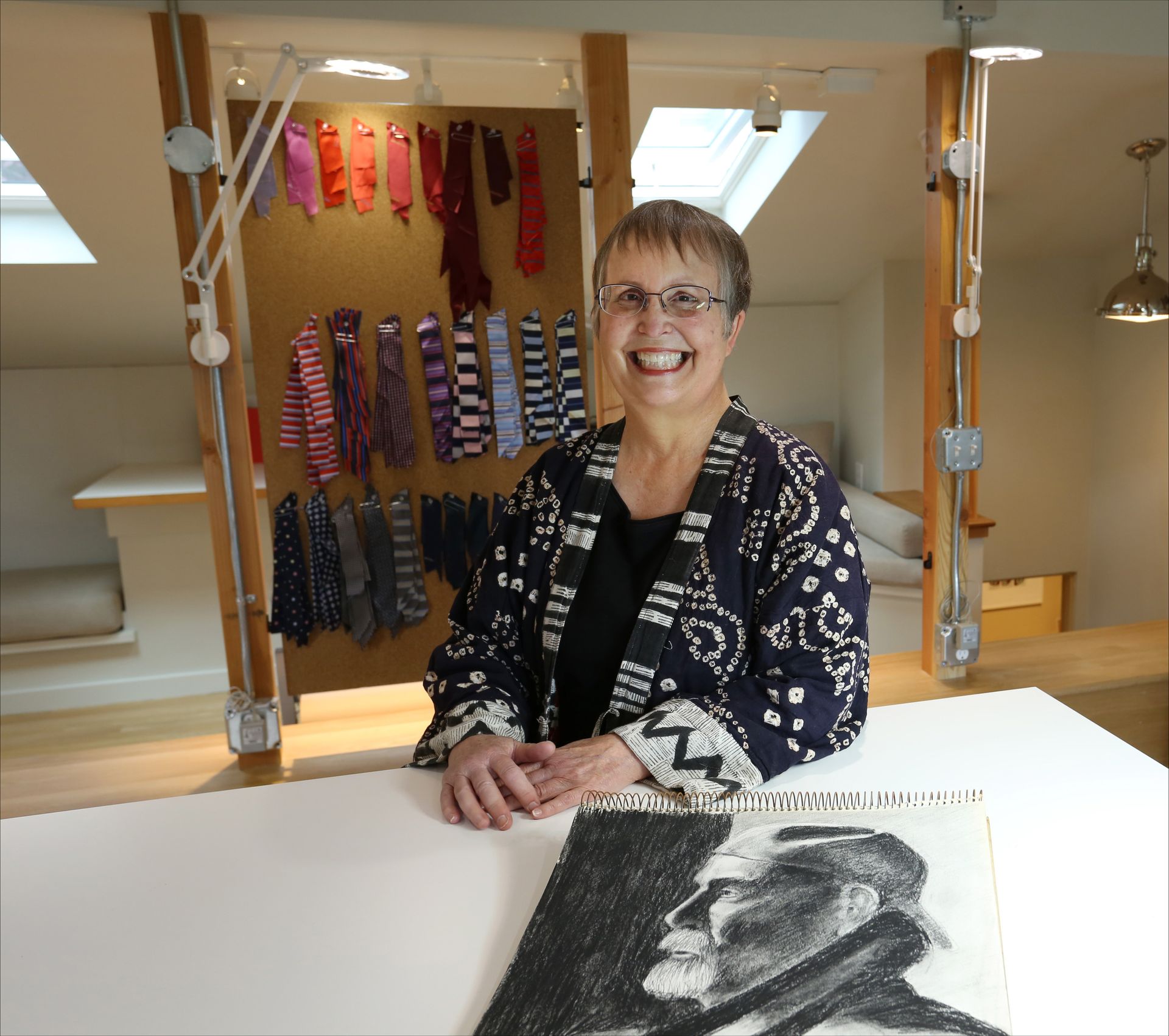 Woman smiling behind a white table in a weaving studio, with hanging textile samples and a black-and-white portrait sketch.